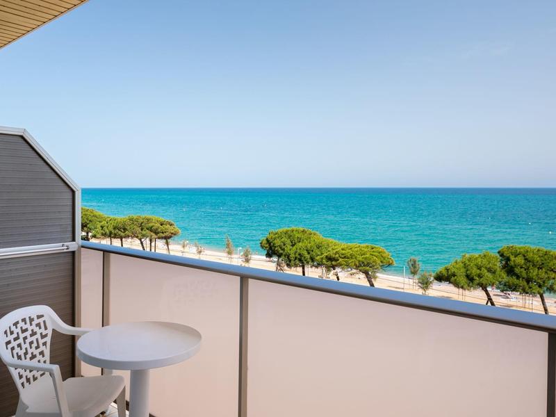 Balcón con mesa y silla blancas, vista al mar y cielo azul claro al fondo.
