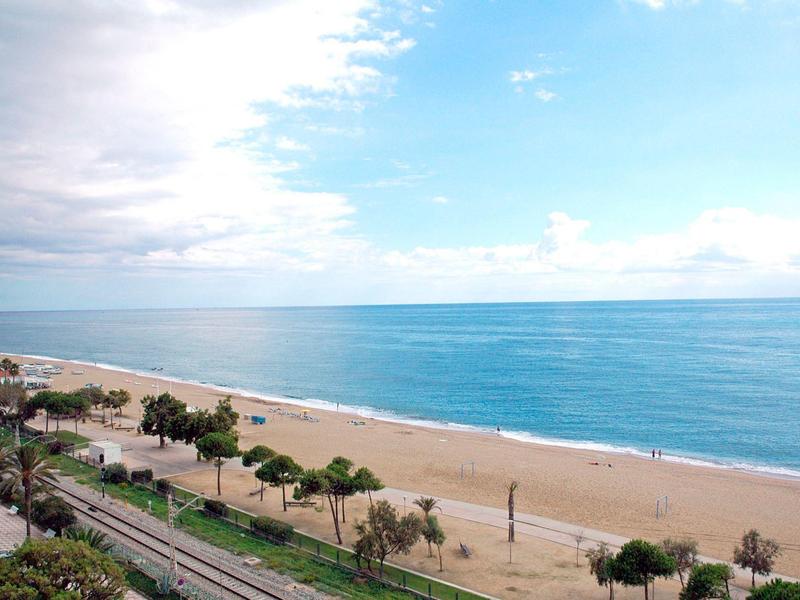 Amplia playa de arena con mar tranquilo y cielo nublado, junto a un paseo con árboles.