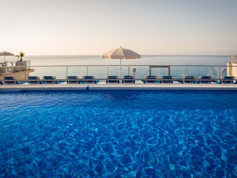 Piscina azul con tumbonas y vista al mar y atardecer.