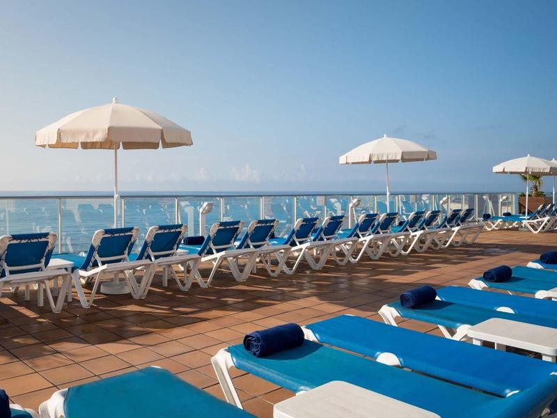 Terraza soleada con tumbonas azules y sombrillas blancas con vista al mar.