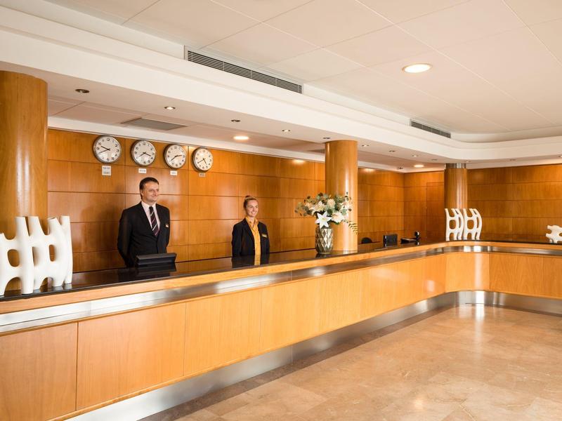 Recepción de hotel con dos empleados detrás del mostrador y relojes mundiales en la pared.