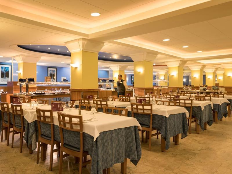 Gran comedor con muchas mesas preparadas y sillas de madera bajo una luz cálida.