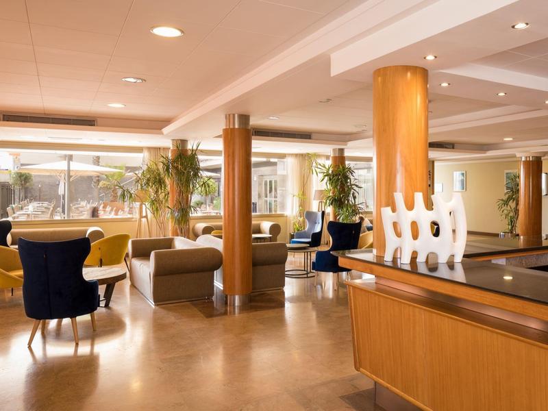 Luminosa y moderna recepción de hotel con áreas de descanso, columnas de madera y grandes ventanas.