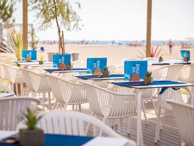 Leere weiße Stühle mit blauen Tischtüchern in einem sonnigen Strand-Café mit Meerblick.