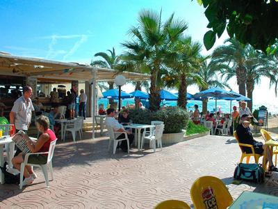 Outdoor-Café mit Tischen, Stühlen, Palmen und Menschen bei sonnigem Wetter.