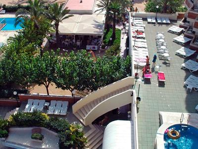 Blick von oben auf Poolanlage mit blauen Pools, Liegestühlen, Palmen und Strand im Hintergrund.