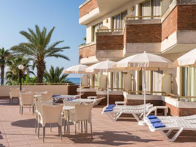 Terrasse mit Sonnenliegen und Tischen vor einem Hotel mit Blick auf Palmen und das Meer.