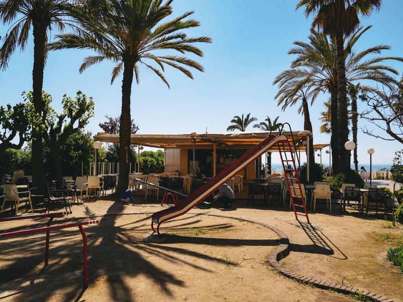 Spielplatz mit Schaukel unter Palmen bei sonnigem Himmel und Sandboden.