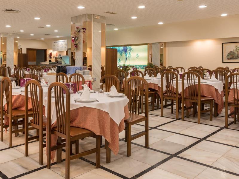 Restaurant mit vielen Holztischen und -stühlen, weißen und braunen Tischdecken, heller Beleuchtung.