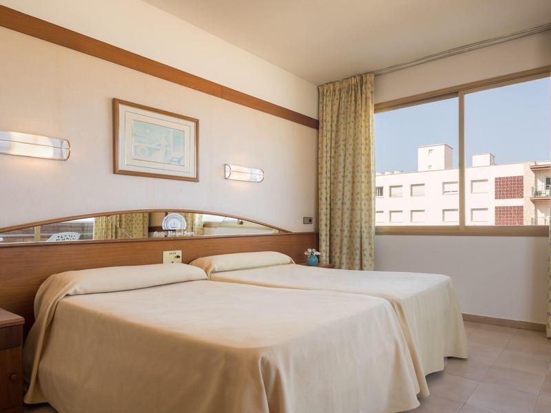 Zweibettzimmer mit weißer Bettdecke, großem Fenster, Vorhängen und Wandschmuck in hellem Raum.