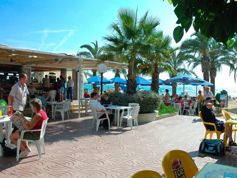 Outdoor-Café mit Tischen, Stühlen, Palmen und Menschen bei sonnigem Wetter.
