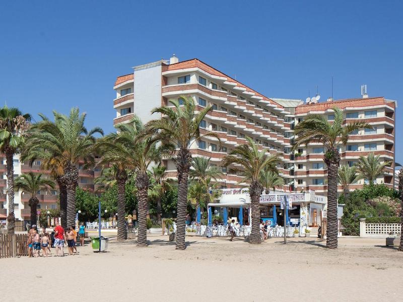 Mehrstöckiges Hotel mit Balkonen hinter Palmen an einem sonnigen Sandstrand.