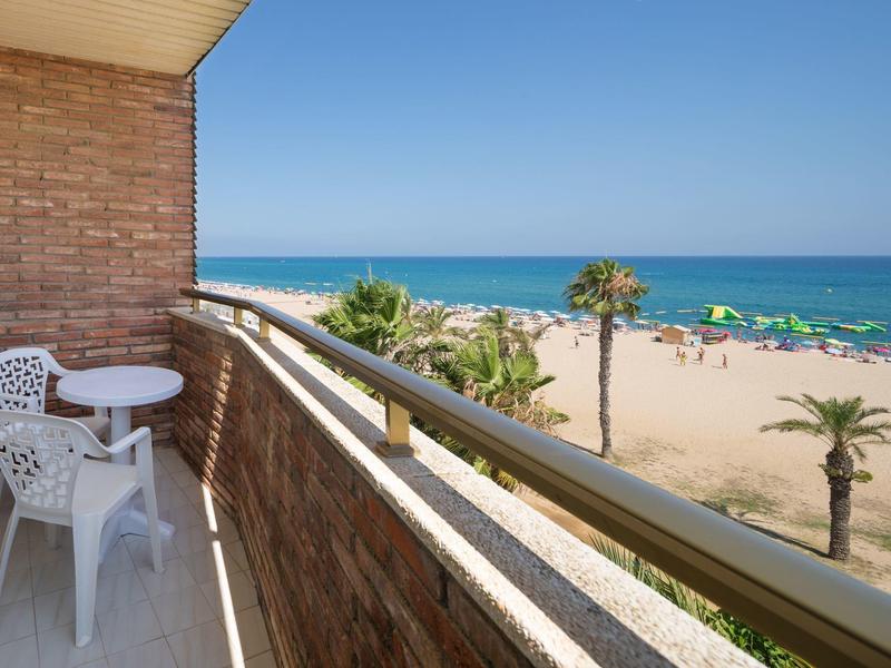 Balkon mit weißen Stühlen und Tisch, Blick auf Sandstrand, Palmen und blaues Meer unter klarem Himmel.