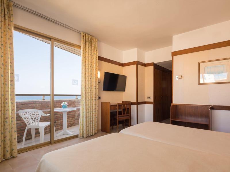 Helles Hotelzimmer mit Doppelbett, Balkon, Fernseher und kleinen Schreibtisch mit Stuhl.