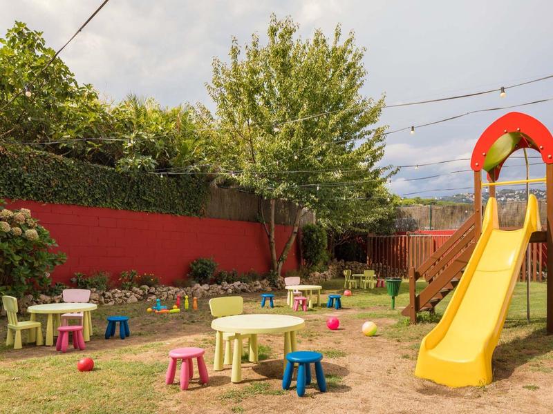 Área de juegos infantiles con tobogán amarillo, mesas y sillas coloridas sobre suelo arenoso, pared roja de fondo