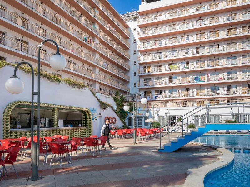 Gran hotel con piscina exterior y sillas rojas bajo cielo azul y sol.