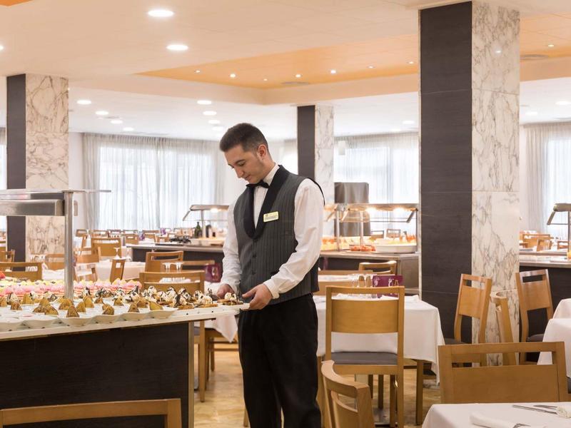 Hombre con chaleco negro arregla comida en un restaurante de hotel luminoso con sillas y mesas de madera.