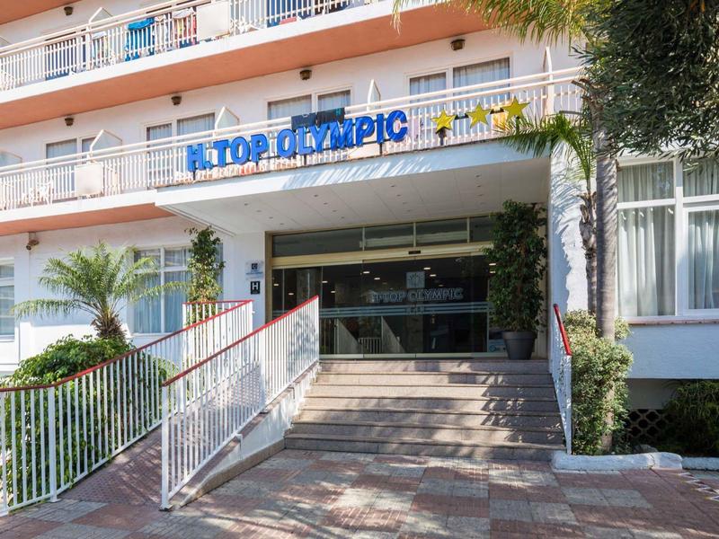 Entrada de un hotel con escaleras, barandales y plantas verdes a los lados.