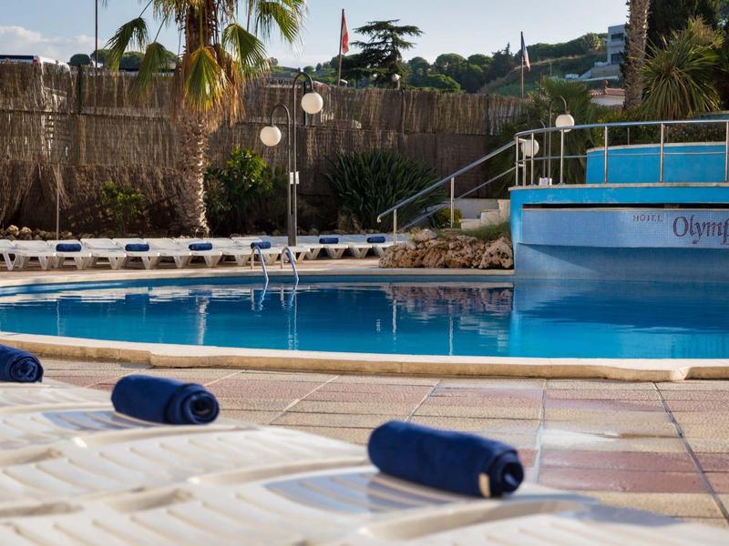Área de piscina vacía con tumbonas blancas y toallas azules bajo palmeras.