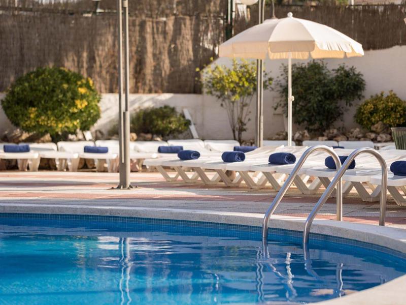 Piscina azul con tumbonas blancas y sombrilla en un jardín de hotel.
