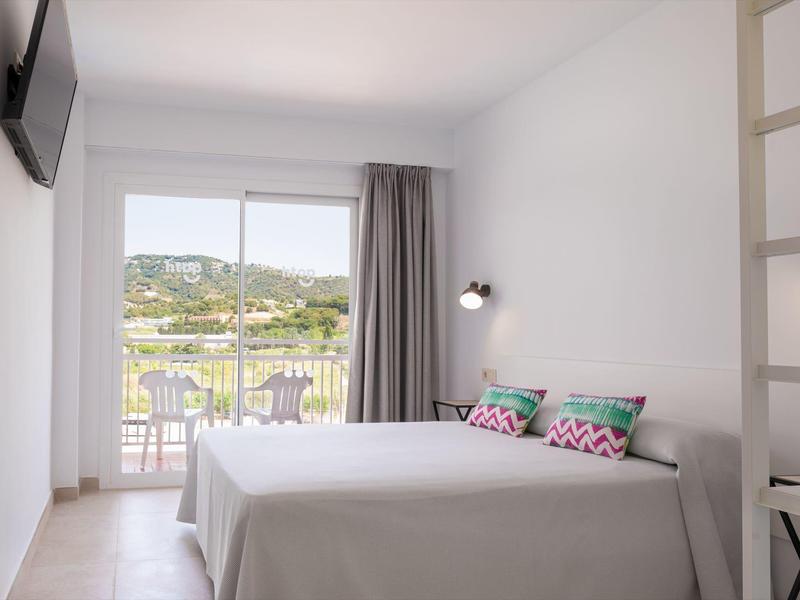 Habitación de hotel luminosa con cama grande, balcón y vistas a colinas verdes.