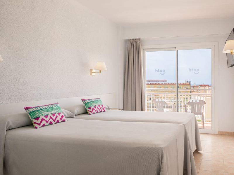 Habitación de hotel luminosa con dos camas individuales, cojines coloridos y balcón con vista al mar.