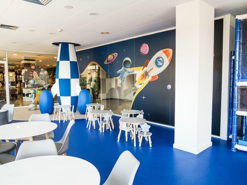 Área infantil con decoración espacial, mesas y sillas blancas en sala azul y blanca.