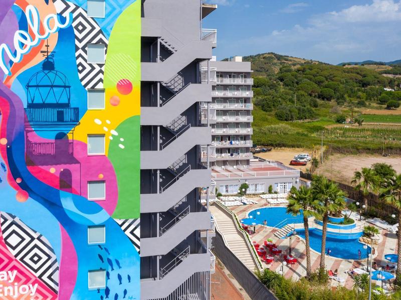Edificio de hotel colorido con piscina y paisaje verde al fondo bajo cielo despejado.