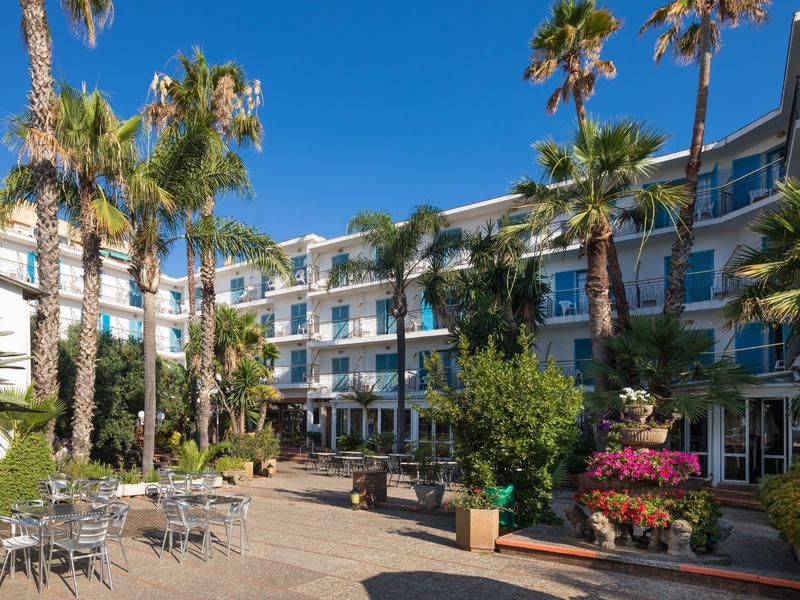 Área exterior de un hotel con palmeras, asientos y flores bajo un cielo azul claro.