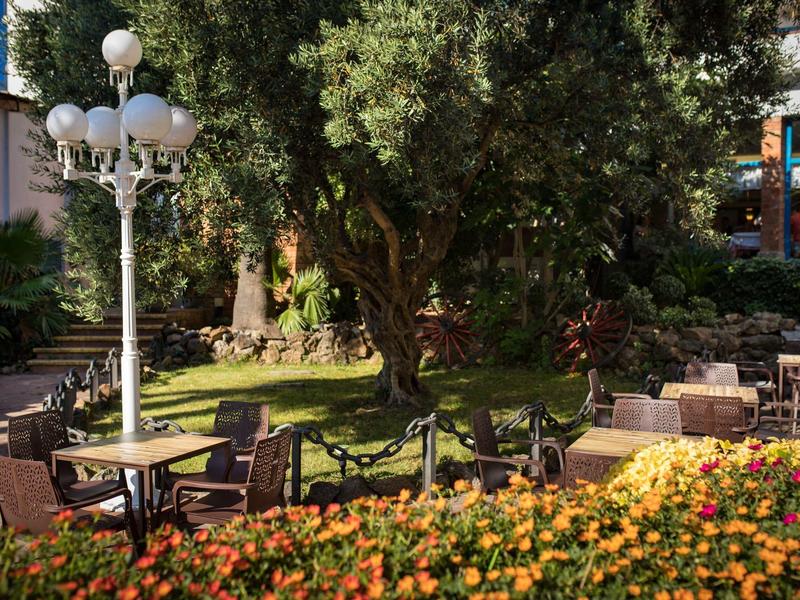 Jardín de hotel con mesas, sillas, flores y un farol blanco en un entorno arbolado.