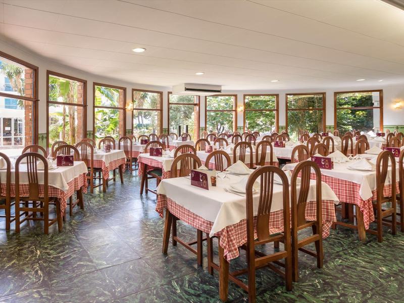 Restaurante luminoso con muchas sillas de madera y mesas cuadradas frente a grandes ventanales.