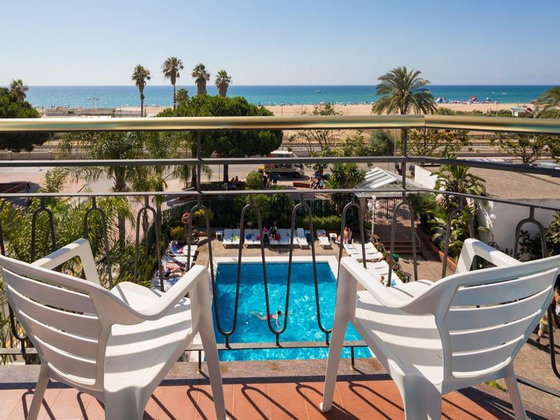 Vista desde el balcón con sillas blancas hacia la piscina, palmeras y el mar en el horizonte con cielo despejado.