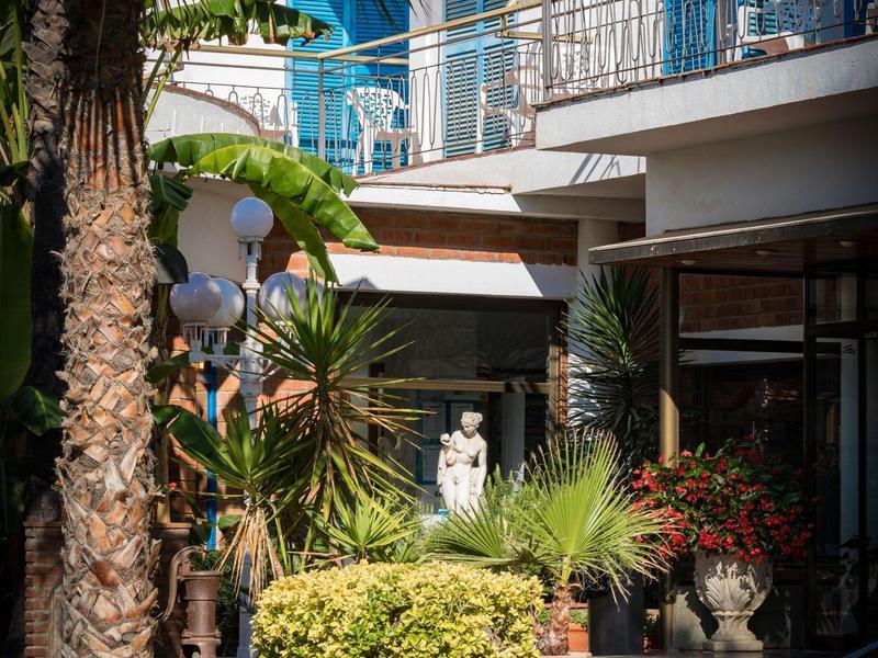 Terraza de hotel con sillas, mesas, palmeras y plantas en flor bajo balcones.