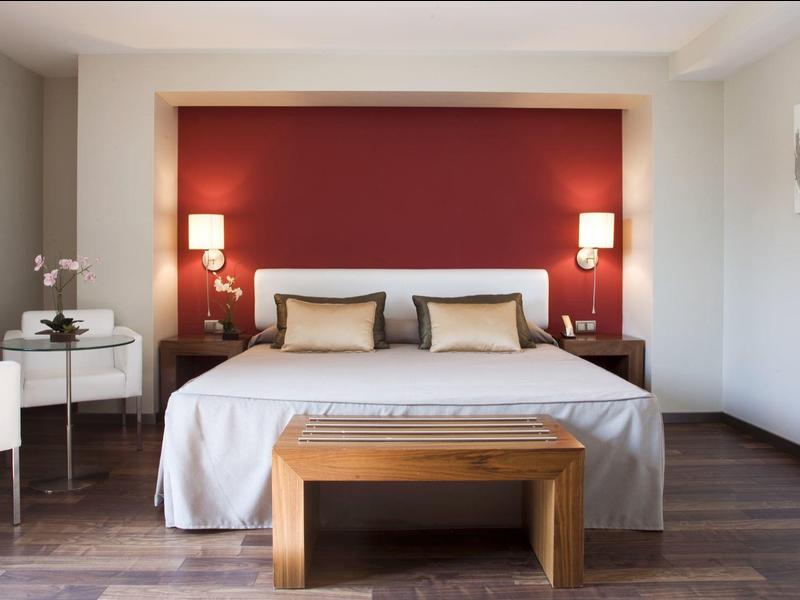 Modern hotel room with large bed, red accent wall, and wooden floor.