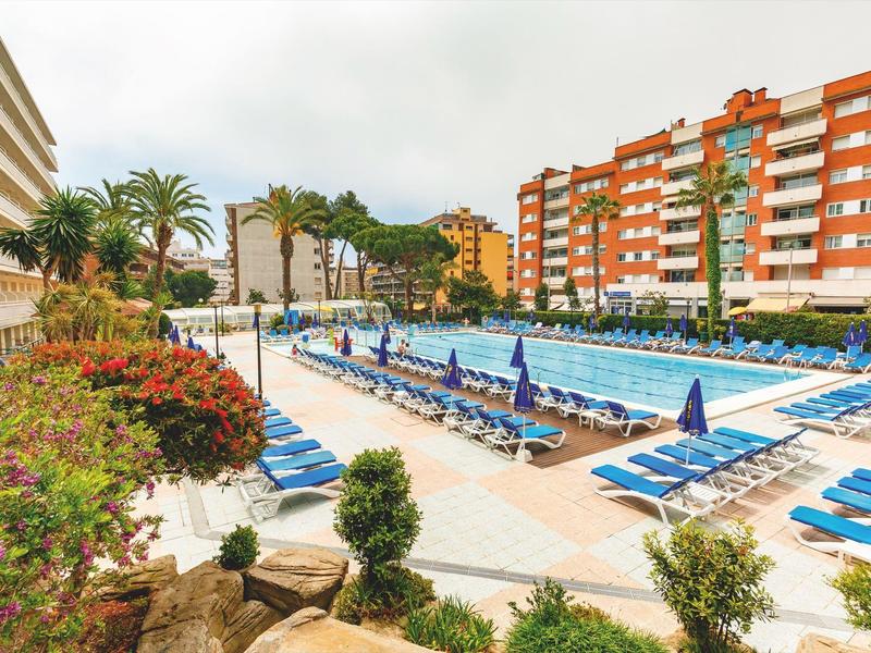 Gran piscina al aire libre con tumbonas azules y sombrillas entre edificios y palmeras.