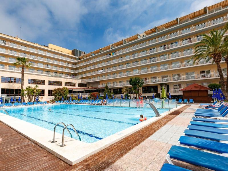 Gran piscina exterior en un hotel con tumbonas azules y palmeras bajo un cielo despejado.
