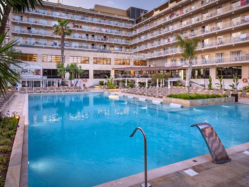 Gran piscina azul de hotel con tumbonas y palmeras en el patio interior de un edificio de varios pisos.