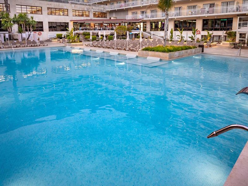 Gran piscina clara del hotel con fuentes de agua y edificio de varios pisos al fondo.