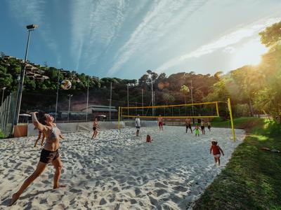 Menschen spielen Volleyball auf einem Sandfeld mit umgebenden Bäumen und Sonnenlicht.