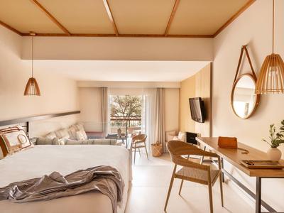 Bright hotel room with two beds, desk, and balcony overlooking palm trees.