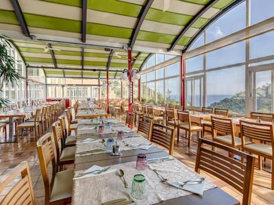 Bright hotel restaurant with large windows, wooden chairs, and green roofs overlooking mountains.