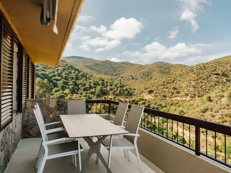 Balcony with table and six chairs overlooking hilly landscape under partly cloudy sky.