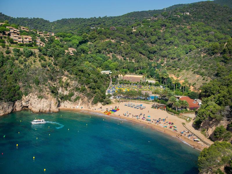 Small sandy cove with clear blue water, surrounded by wooded hills with sun loungers and boats.