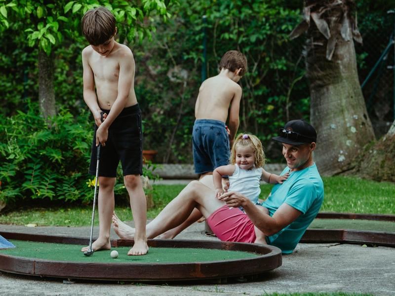 Children and an adult playing mini-golf in a green, wooded area.