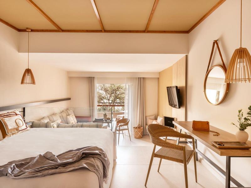 Bright hotel room with two beds, desk, and balcony overlooking palm trees.