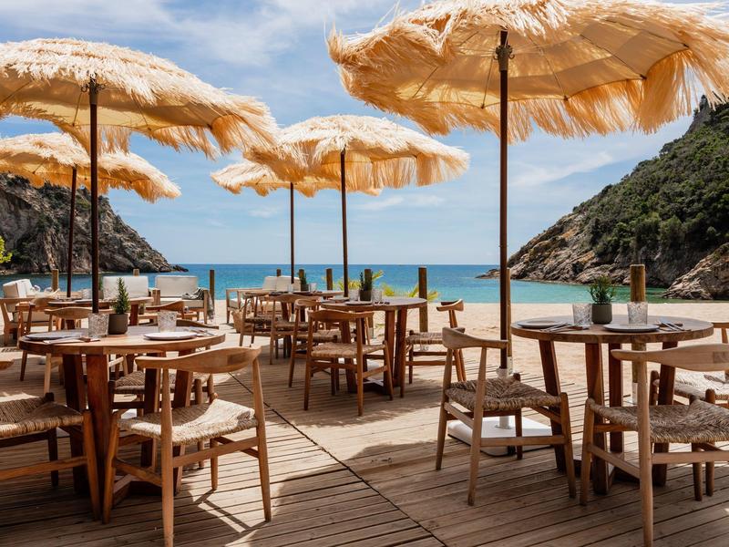 Empty beach area with wooden tables, chairs, and straw umbrellas overlooking the sea.