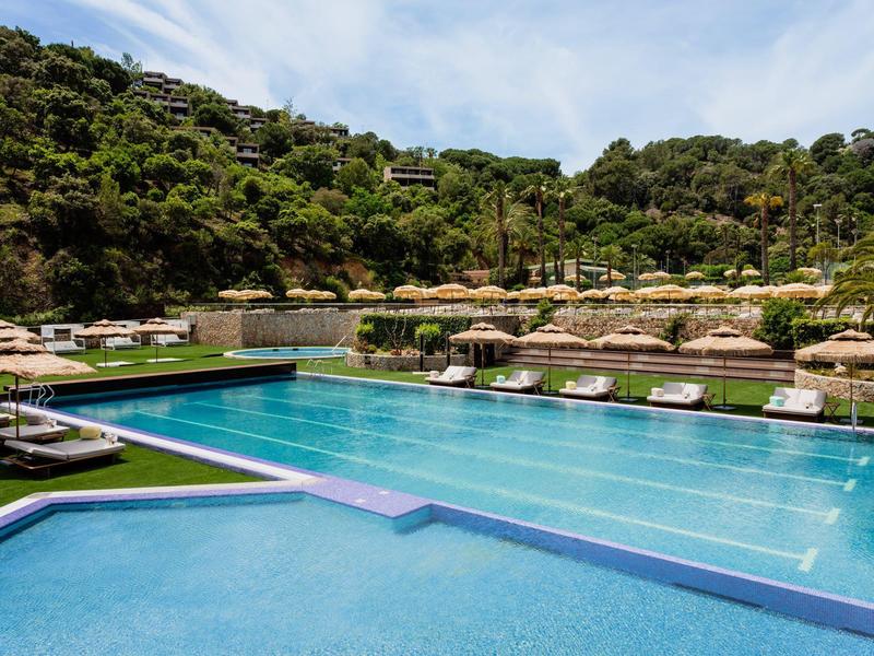 Large hotel pool with blue water, surrounded by sun loungers and green hills.