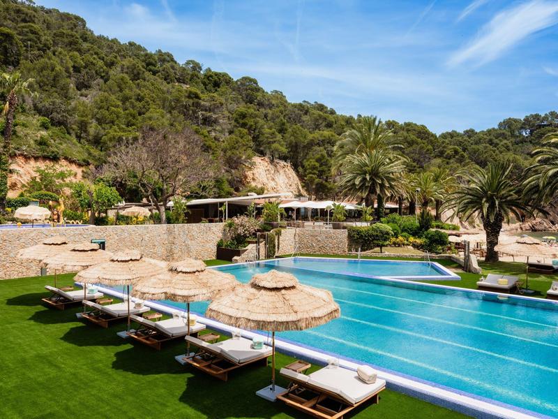 Large pool with sunbeds and umbrellas in front of wooded hills under blue sky.
