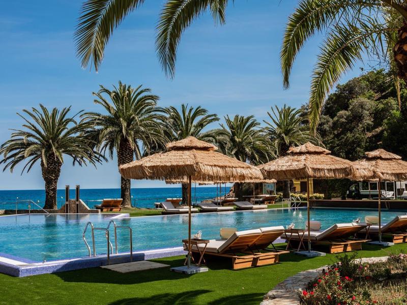 Luxurious pool area with sun loungers, palm trees, and sea view under a blue sky.