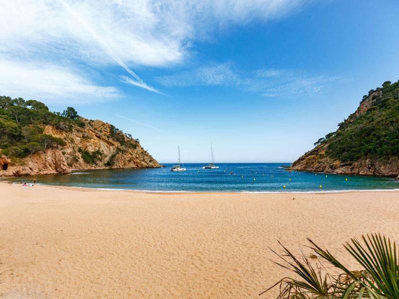 A quiet sandy beach between two wooded cliffs with sailboats in the bay.